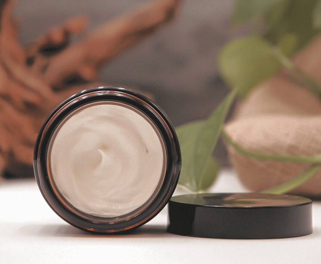 Un pot en verre brun rempli de crème blanche est posé sur une surface dont le couvercle noir a été retiré, tandis que de la verdure et un objet en bois sont flous à l'arrière-plan.