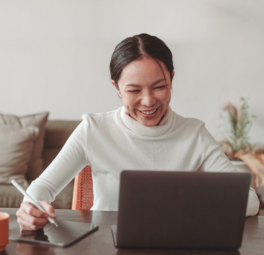 Une femme en col roulé blanc est assise à une table, souriant tout en utilisant un ordinateur portable et en écrivant sur une tablette à l'aide d'un stylet. Il y a un canapé et des plantes à l'arrière-plan.