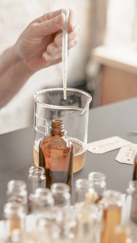 Une personne utilise un compte-gouttes pour transférer un liquide dans un bécher rempli d'un liquide ambré, entouré de petites bouteilles en verre et d'étiquettes sur une table de laboratoire.